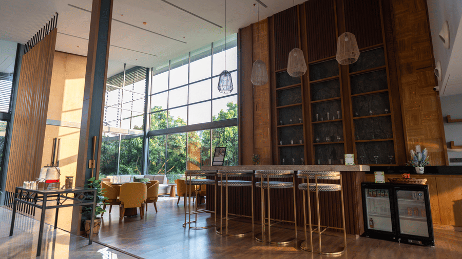 A sophisticated bar area at Wyndham Garden featuring a long counter with high stools, a large built-in wooden shelving unit, and natural light streaming through floor-to-ceiling windows with outdoor views.
