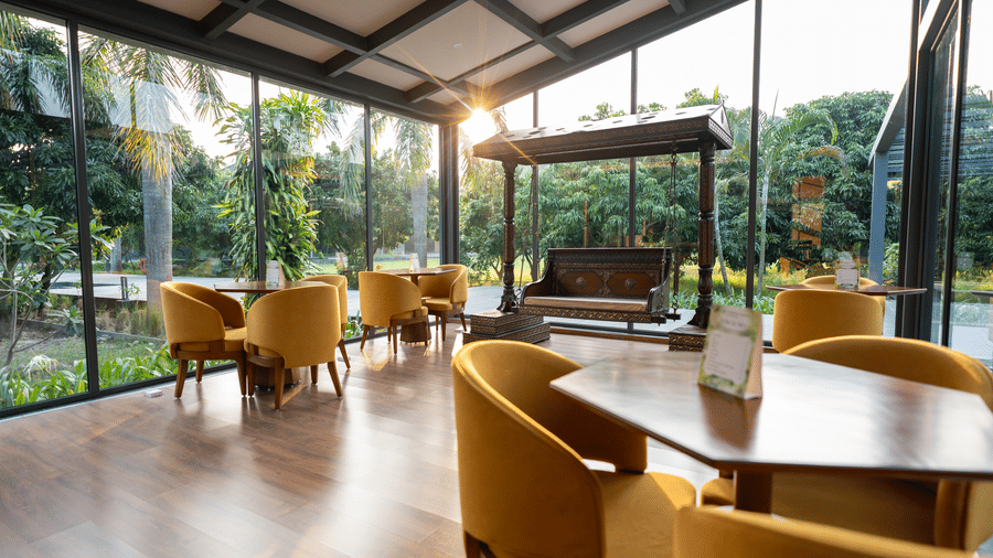 A bright glass-walled lobby at Wyndham Garden with a wooden floor, round tables with upholstered armchairs, a central wooden swing bench, and views of lush greenery outside.