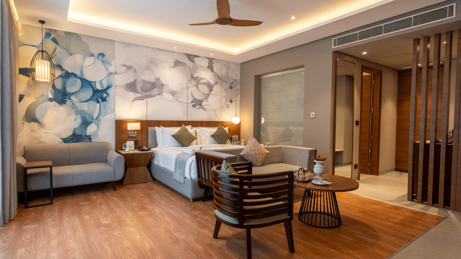 A modern bedroom at Wyndham Garden featuring 2 large beds, a round wooden central table, a seating area with chairs, and heavy curtains covering the window wall.