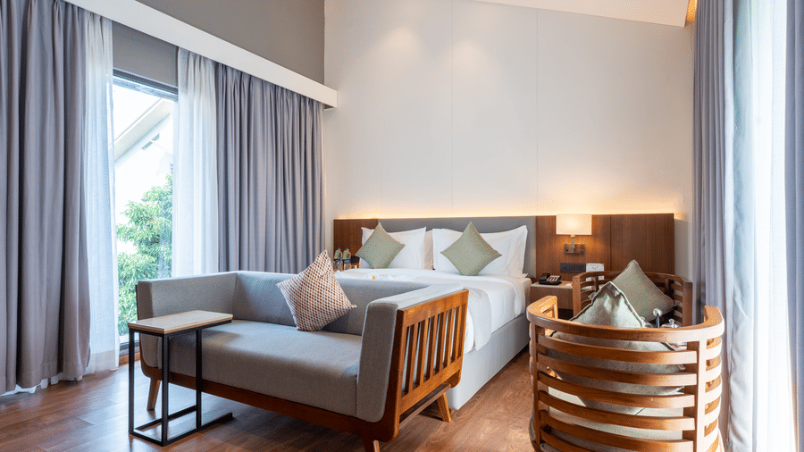 A Wyndham Garden bedroom with a large bed, a wooden lounge chair, and a relaxing sitting area by sliding glass doors with curtains.