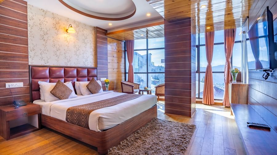 A stylish hotel room with a large bed, TV, and large windows with a view at the Yashshree Jai Hotel & Banquet, Darjeeling.