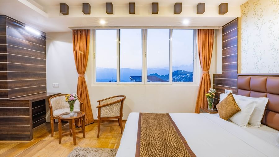 A spacious hotel bedroom with a large bed, seating area, and a large window overlooking the town at the Yashshree Jai Hotel & Banquet, Darjeeling.