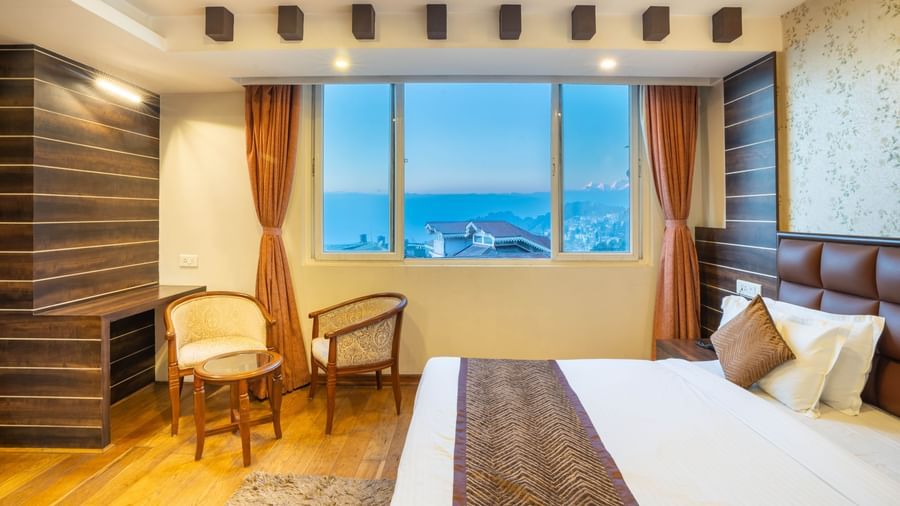 A modern hotel bedroom with a large bed, seating area, and a large window overlooking the town at the Yashshree Jai Hotel & Banquet, Darjeeling.