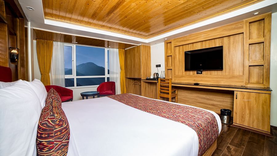 Modern hotel room with a large bed dressed in white linens and red decorative accents, wooden ceiling and flooring, a flat-screen TV mounted on a wooden entertainment unit, and a window offering scenic mountain views with two red chairs and a small table n