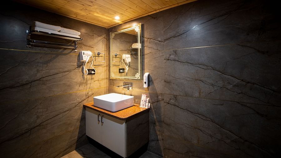 Modern bathroom with dark marble-patterned walls and a wooden ceiling, featuring a countertop sink with a mirror above it, wall-mounted hair dryer, soap dispensers, and soft warm lighting - Yashshree Lachen.