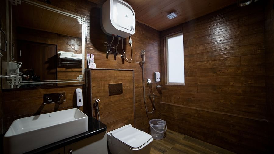 Contemporary bathroom with dark wooden walls and flooring, featuring a square countertop sink with mirror, wall-mounted toilet, overhead water heater, a shower area with handheld fixtures, and a plastic bucket on the floor - Yashshree Lachen.