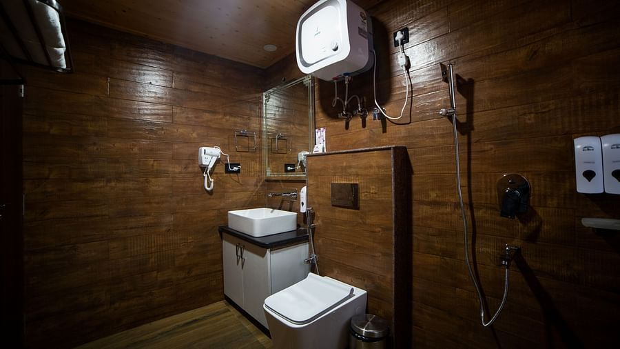 Modern bathroom with dark wood-panelled walls, featuring a square countertop sink with mirror, wall-mounted toilet, water heater, handheld shower fixtures, and mounted dispensers for toiletries - Yashshree Lachen.