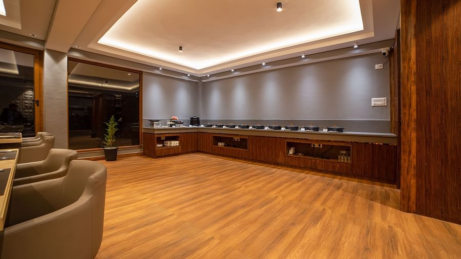 A dining area with wooden flooring, a long U-shaped counter against grey walls, recessed ceiling lighting, a few cushioned chairs on the left, and a potted plant near the glass window - Yashshree Pelling Chekhim Retreat.