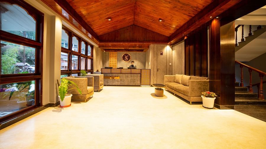 Spacious hotel lobby with a wooden vaulted ceiling, warm lighting, large windows, neutral-toned sofas, potted plants, and a reception desk in the background - Yashshree Pelling Chekhim Retreat.