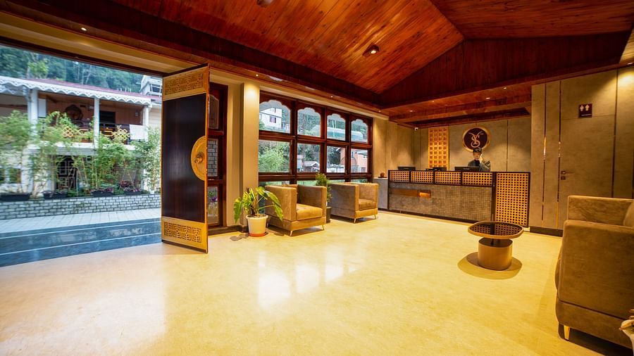 Bright hotel lobby with wooden vaulted ceiling, open entrance leading to a garden area, cosy seating with potted plants, and a reception desk in the background - Yashshree Pelling Chekhim Retreat.