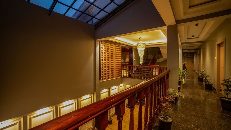 Indoor hallway with a wooden railing overlooking a lower level, a slanted glass ceiling, recessed wall lighting, potted plants along the corridor, and doorways leading to rooms - Yashshree Pelling Chekhim Retreat.