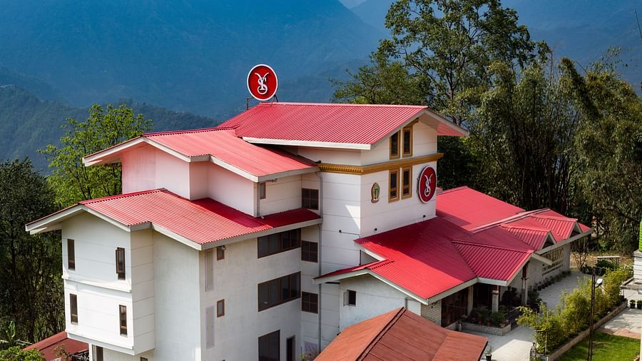 Scenic view of a multi-story white building with red roofs, surrounded by lush greenery and mountains in the background under a clear sky - Yashshree Pelling Chekhim Retreat.