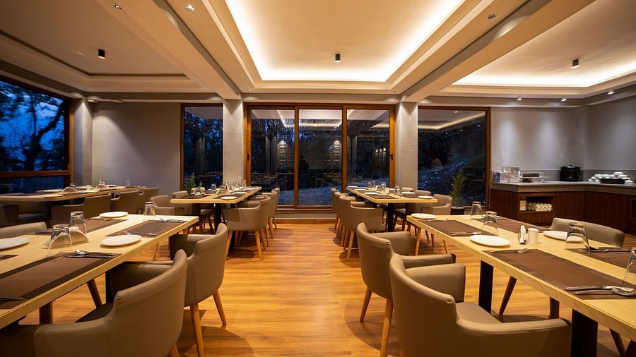 Dining area with wooden flooring, multiple rectangular tables set with plates and glasses, cushioned chairs, recessed ceiling lighting, and large glass windows and doors overlooking trees outside - Yashshree Pelling Chekhim Retreat.