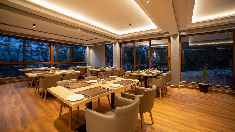 Modern, warmly lit restaurant interior with wooden floors, beige chairs, and tables set for dining, surrounded by large windows showing a view of trees at dusk - Yashshree Pelling Chekhim Retreat.