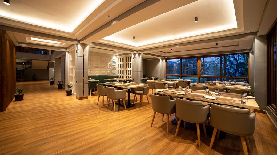 A large room with wooden flooring, several rectangular dining tables surrounded by cushioned chairs, a ceiling with recessed lighting panels, large windows on one side, and some potted plants placed along the walls - Yashshree Pelling Chekhim Retreat.