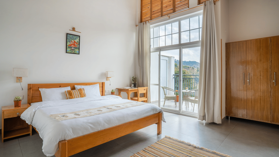 A wide shot of a Ziran Retreat bedroom with a large bed, a wooden bedside table, and large glass doors open to a balcony with a scenic view.