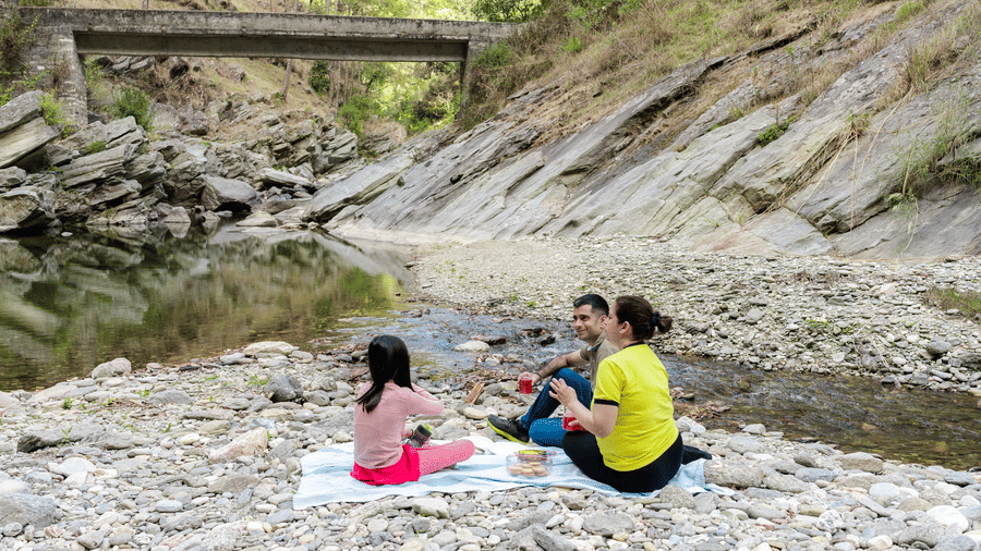 A woman in a yellow shirt and a child in a pink shirt are sitting on a blanket near a river at Ziran Retreat.