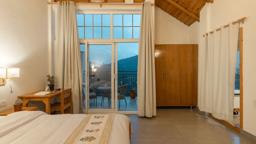 A close-up of a Ziran Retreat bedroom showing a bed with a white comforter, a bedside table with a lamp, and large glass doors leading to a balcony.