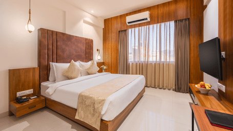 A comfortable king-size bed with soft pillows and a decorative throw faces a mounted television in a well-lit hotel room with a window - Lords Eco Inn Ambaji.