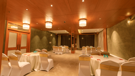 A warmly lit banquet hall at Lords Eco Inn Apple Gate is set with round tables draped in white, surrounded by chairs with gold sashes.