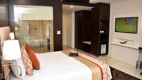 A bedroom in Lords Plaza Surat featuring a double bed, plush pillows and a glass panelled washroom with half-pulled blinds