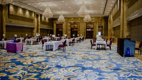 A large, empty banquet hall featuring patterned blue and gold carpets, chandeliers, and round tables set up for an event.