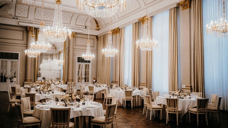 A luxurious ballroom with round tables, white linens, and ornate crystal chandeliers hanging from a high ceiling.