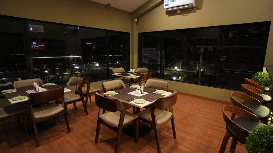 Cosy indoor dining area with modern wooden tables and cushioned chairs, large glass windows offering night views, and bar stools lined along a dark counter - Gaurav Lords Resort Shrivardhan