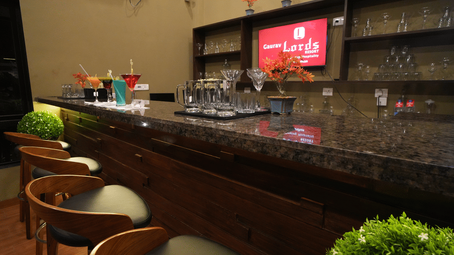 Close-up view of a modern bar setup with wooden high chairs, granite countertop, glassware, vibrant cocktails, decorative plants, and neatly arranged shelves in the background - Gaurav Lords Resort Shrivardhan