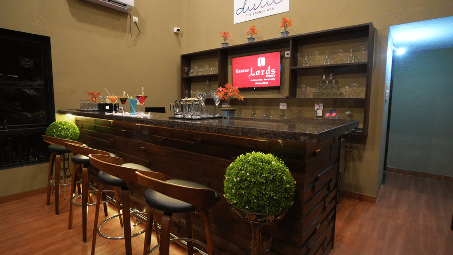 Modern indoor lounge bar with a sleek wooden counter, stylish high chairs, decorative plants, glass shelves, and a variety of colourful cocktails on display - Gaurav Lords Resort Shrivardhan
