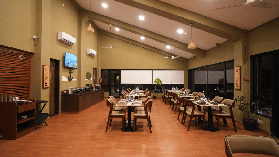 A restaurant with set tables and chairs, featuring a high, sloped ceiling and warm lighting. It includes a buffet area, air-conditioning units, and a wooden floor - Gaurav Lords Resort Shrivardhan.
