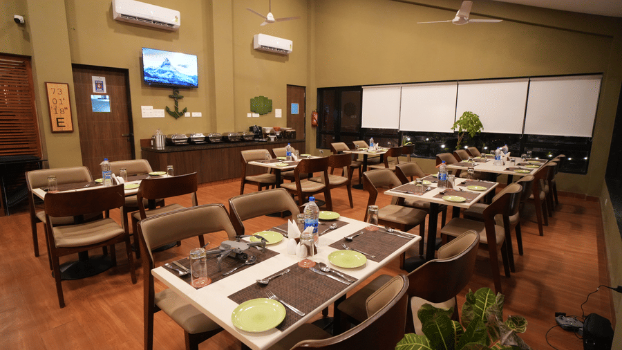 A restaurant with neatly arranged tables and chairs, offering a warm and inviting, dimly lit atmosphere. It features a television on the wall, air-conditioning units, and potted plants for added charm - Gaurav Lords Resort Shrivardhan.