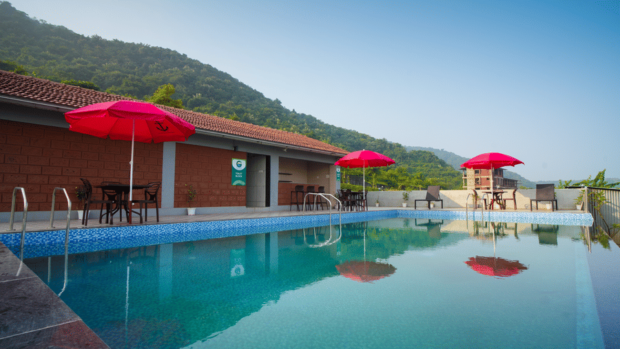 A swimming pool with red umbrellas and lounge chairs, set against a backdrop of mountains and a building - Gaurav Lords Resort Shrivardhan.