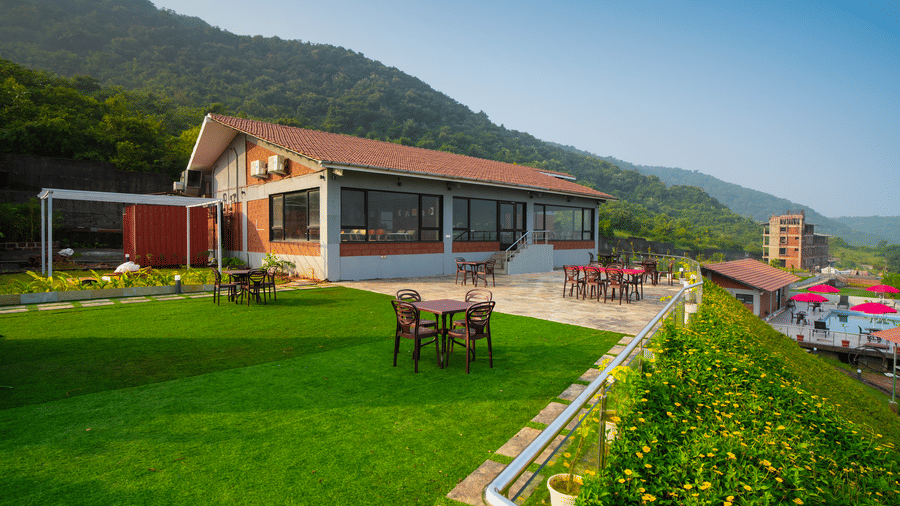 A building with a red roof and large windows, surrounded by a green lawn and a mountain backdrop, featuring outdoor tables and chairs and a metal railing in the foreground - Gaurav Lords Resort Shrivardhan.