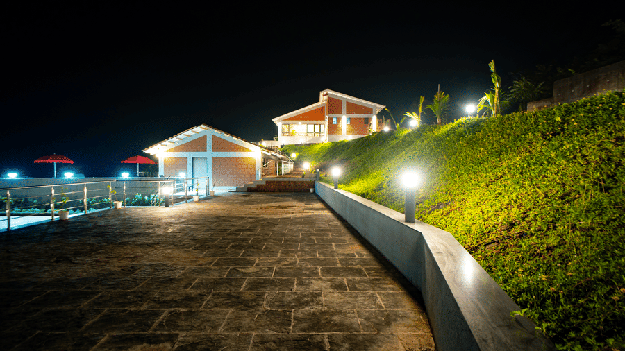 A stone pathway leading to illuminated buildings at night, with a green hillside on the right - Gaurav Lords Resort Shrivardhan.
