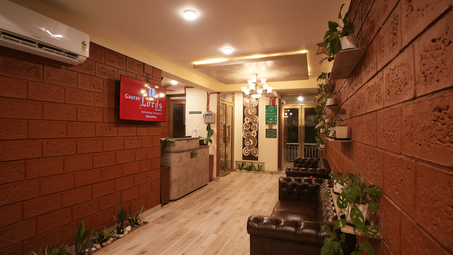Interior hallway of a resort with textured red brick walls, a wooden reception desk, a dark leather sofa, and potted plants. A television is mounted on the wall - Gaurav Lords Resort Shrivardhan.