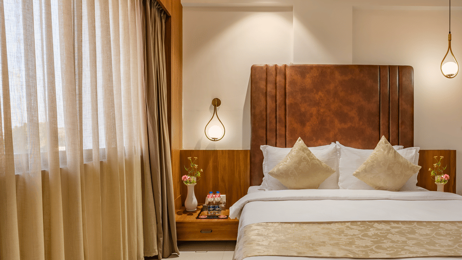 A comfortable bed is situated between two nightstands with lamps in a hotel room with layered window coverings - Lords Eco Inn Ambaji.