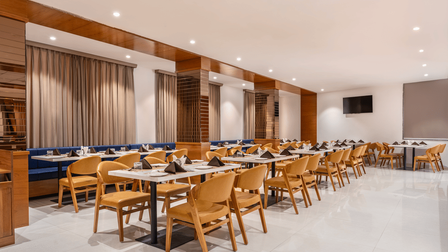 A brightly lit dining area features numerous set tables with chairs and booth seating along the wall - Lords Eco Inn Ambaji.
