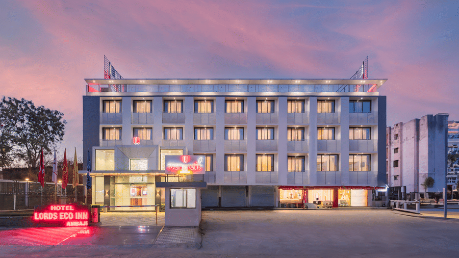 A modern hotel building with many windows and a bright red sign stands beside a paved area under a twilight sky - Lords Eco Inn Ambaji.