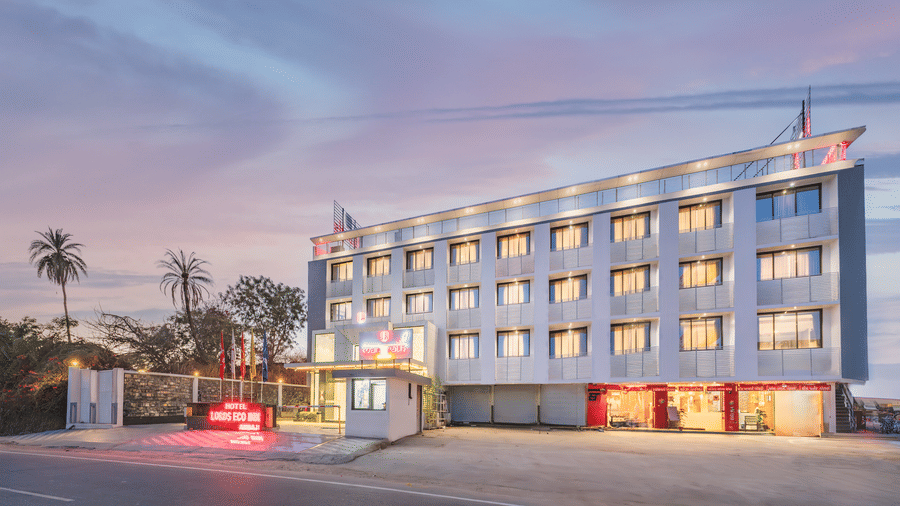 A multi-story building with illuminated windows and a red sign stands beside a paved area at dusk - Lords Eco Inn Ambaji.