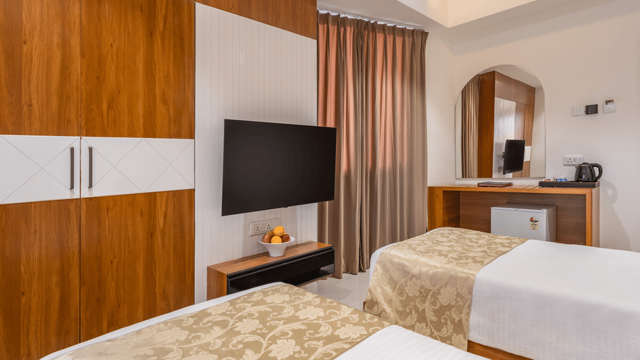 A hotel room features two single beds, a wall-mounted television, a wardrobe, and a desk with a mirror - Lords Eco Inn Ambaji.