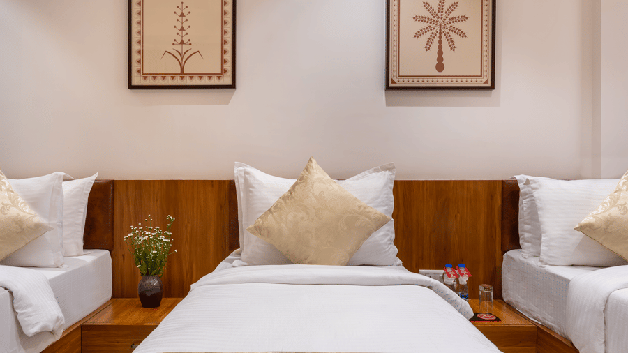 Three single beds with white linens and decorative pillows are positioned under framed botanical prints on a wall - Lords Eco Inn Ambaji.
