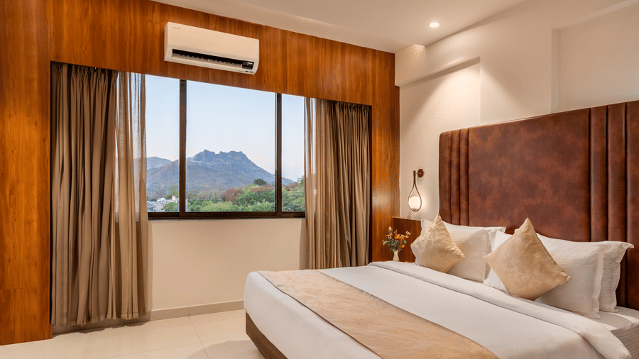 A comfortable king-size bed with soft pillows and a throw faces a window with curtains revealing a mountain view - Lords Eco Inn Ambaji.