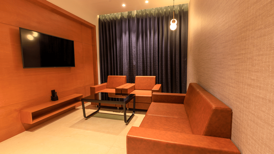A warmly lit lounge space of Executive Room at Lords Eco Inn Apple Gate features orange sofas and armchairs, a black coffee table, and a wall-mounted TV.