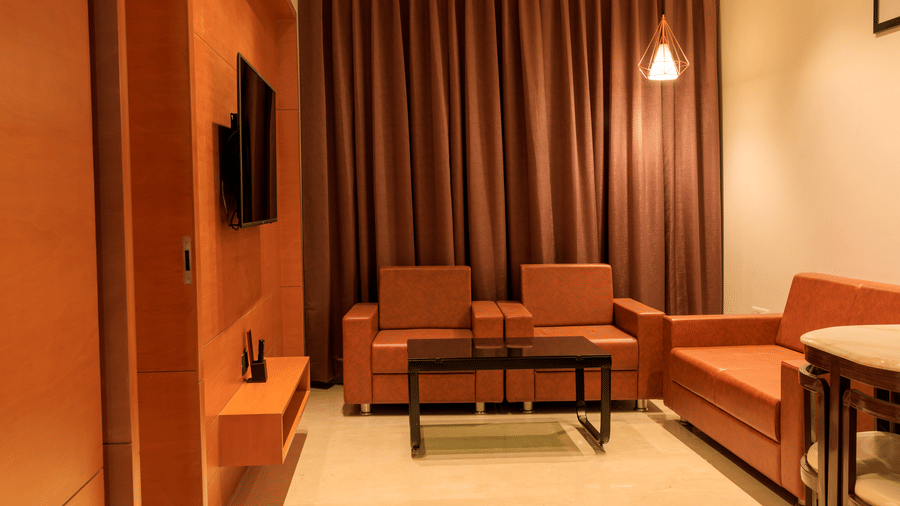 A hotel room at Lords Eco Inn Apple Gate features orange sofas, a black coffee table, a wall-mounted television, and brown curtains.