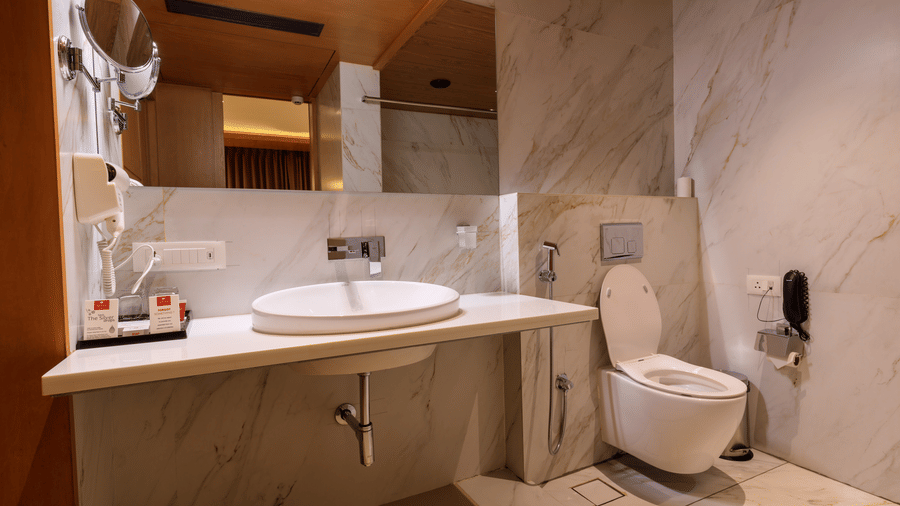 A modern hotel bathroom features a white oval sink on a light countertop, a toilet, a large mirror, and marble-effect wall tiles at Lords Eco Inn Apple Gate