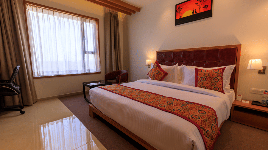 A well-lit hotel room at Lords Eco Inn Apple Gate features a double bed with patterned accents, a desk, a chair, and window curtains.