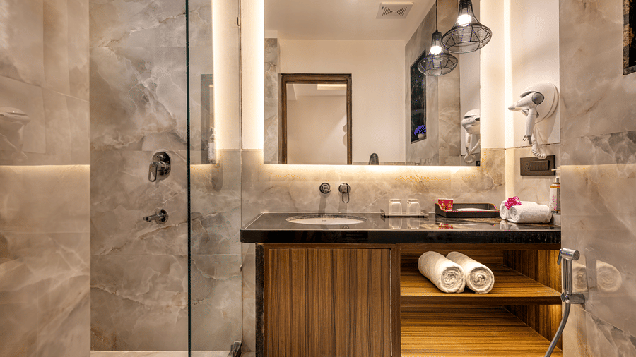 A modern bathroom with a glass-enclosed shower, a wooden cabinet with a black counter top and a sink, a large wall mirror with pendant lights at Lords Eco Inn Dehradun.