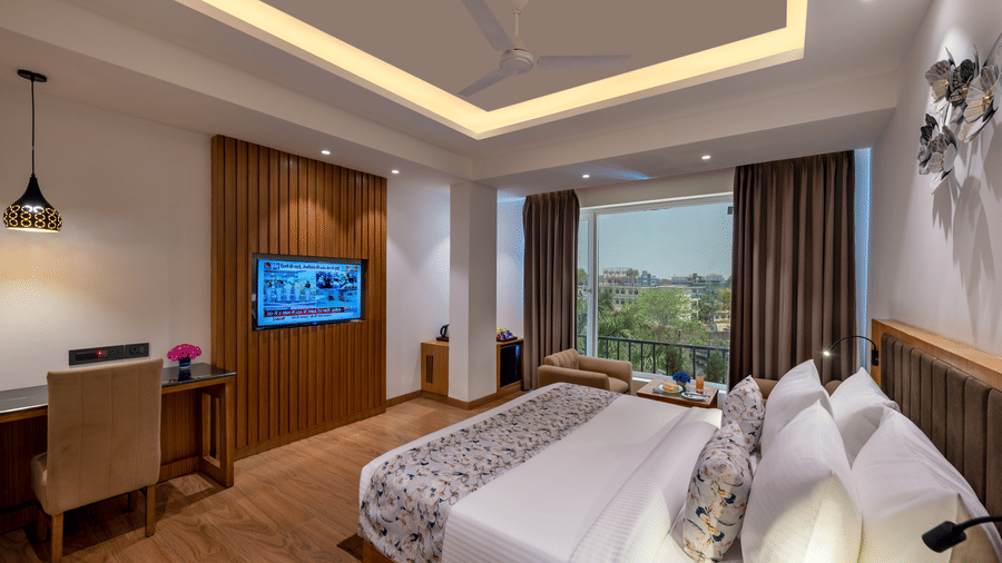 A modern designed room with a king size bed, a table and a chair set, a mounted T.V., a couch on the window side overlooking the greenery and the cityscape at Lords Eco Inn Dehradun.