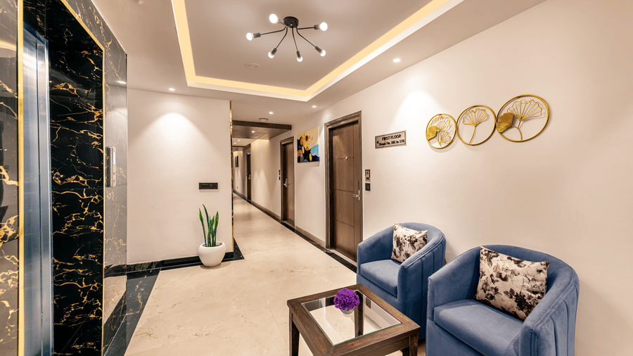 Well-lit hotel corridor with blue armchairs, a wooden table, modern ceiling lights, and an elevator on the left at Lords Eco Inn Dehradun.